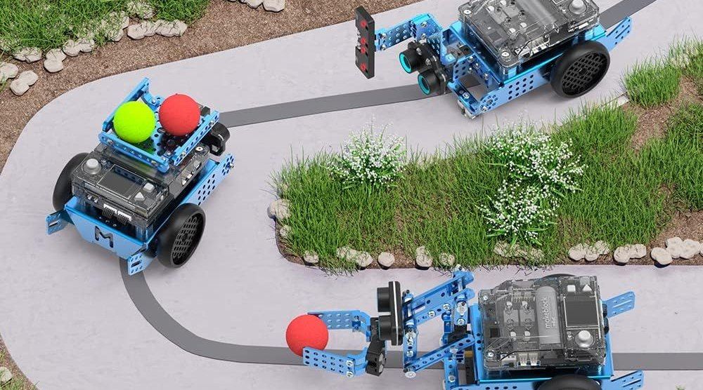 Drei verschieden aufgebaute blaue Roboterfahrzeuge mit Metallstreben fahren auf einem gepflasterten Parcours zwischen Grasflächen, eines transportiert zwei Schaumstoffbälle