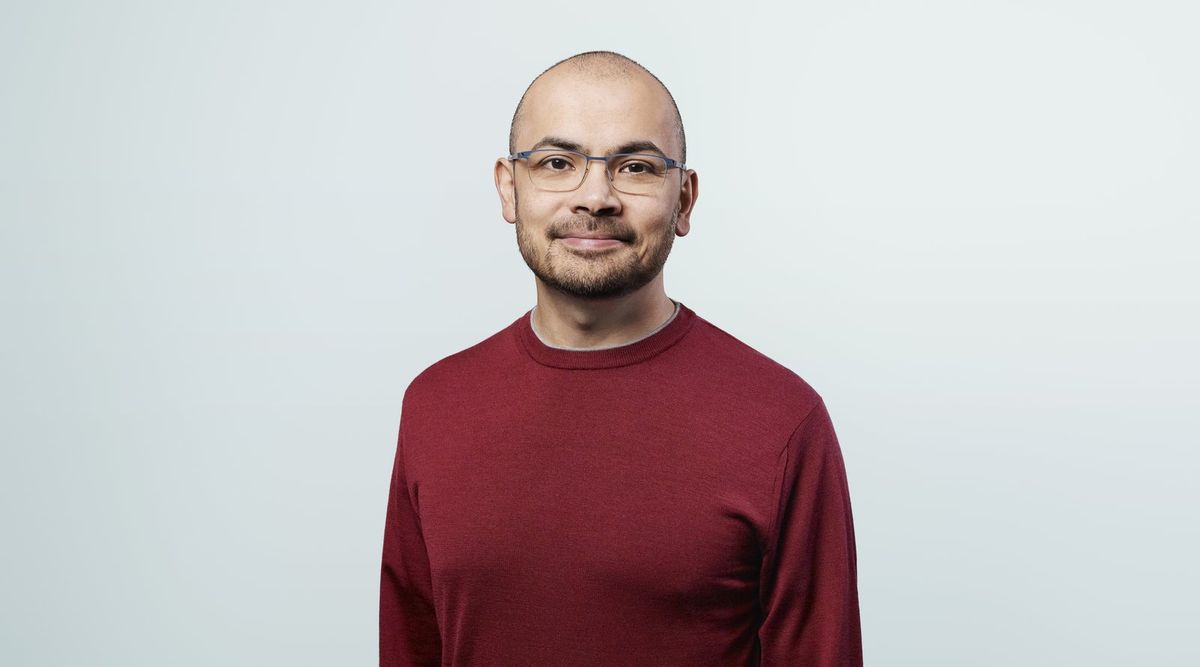 Demis Hassabis in dunkelrotem Pullover mit schmaler Brille und kurzem Bart lächelt leicht in die Kamera, Studioporträt vor hellgrauem Hintergrund